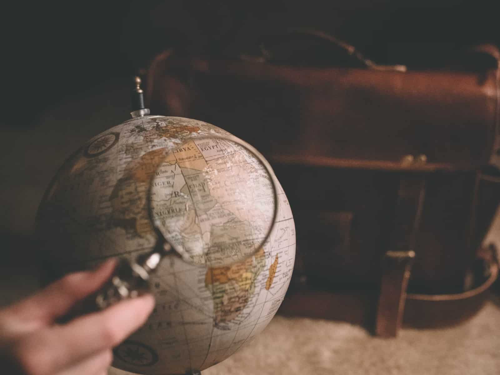 hand holding a magnifying glass over a globe in a room with a leather satchel in the background